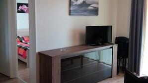 Una sala de estar moderna con un televisor de pantalla plana sobre una consola de madera, un cuadro de una ola en la pared y vistas al exterior a través de una ventana con cortinas.