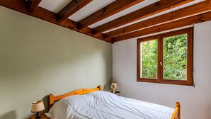 A bedroom with a wooden bed, a nightstand with a lamp, a window with a view of greenery, and a sloped ceiling with wooden beams. - Charming 4-Person Home, Quiet Area Near Beaches, Biscarrosse (Biscarrosse Plage)