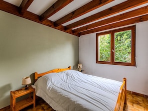 A bedroom with a wooden bed, a nightstand with a lamp, a window with a view of greenery, and a sloped ceiling with wooden beams.