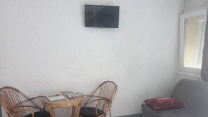 Huoneessa on seinälle asennettu televisio, pyöreä pöytä, jossa on kaksi rottinkituolia, harmaa sohva, jossa on punainen koristetyyny, ja ikkuna, jossa on sälekaihtimet.