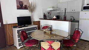 A compact kitchen with white cabinets, a microwave, and a sink. There is a dining table with four chairs, each with a red cushion. A television is placed on a cabinet, and a vase with dried plants is on the table. - Gite l'occitan 5 people (autignac)