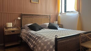 A bedroom with a bed, a nightstand, a lamp, a wicker chair, and a window with curtains. - Gite l'occitan 5 people (autignac)