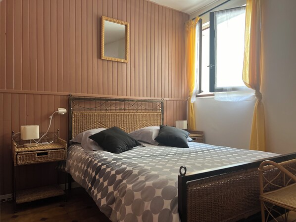 Un dormitorio con cama, mesita de noche, lámpara, silla de mimbre y ventana con cortinas.