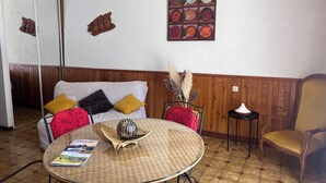 A room with a round table, a sofa with colorful cushions, a wooden panel wall, and a wall art piece. - Gite l'occitan 5 people (autignac)