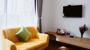 A modern living room with a yellow sofa, a flat-screen TV mounted on the wall, a wooden side table with snacks and drinks, and a window with curtains. - BB's Cozy House & Apartment (Da Nang)