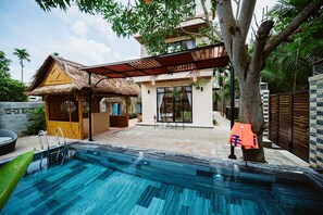 Une piscine avec une terrasse en bois, un abri au toit de chaume et une maison moderne dotée d’un patio couvert.