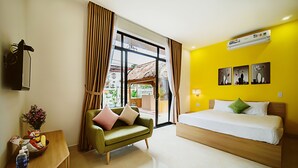 A hotel room with a bed, a green sofa, a small table with a basket of fruits, and a window with curtains. - BB's Cozy House & Apartment (Da Nang)