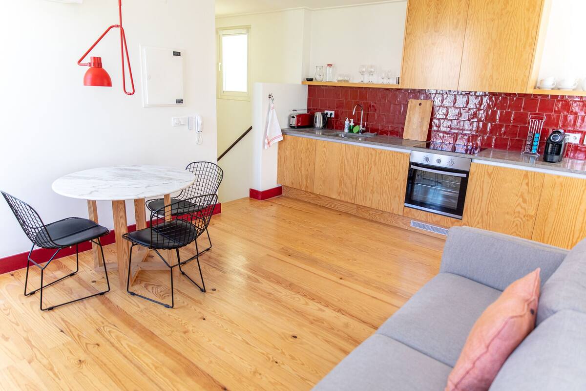 A modern kitchen with wooden cabinets, a red backsplash, and a dining area with a marble table and black wire chairs.