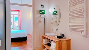 Una habitación con piso de madera, una cama, un baño con espejo, un radiador y un estante con toallas.