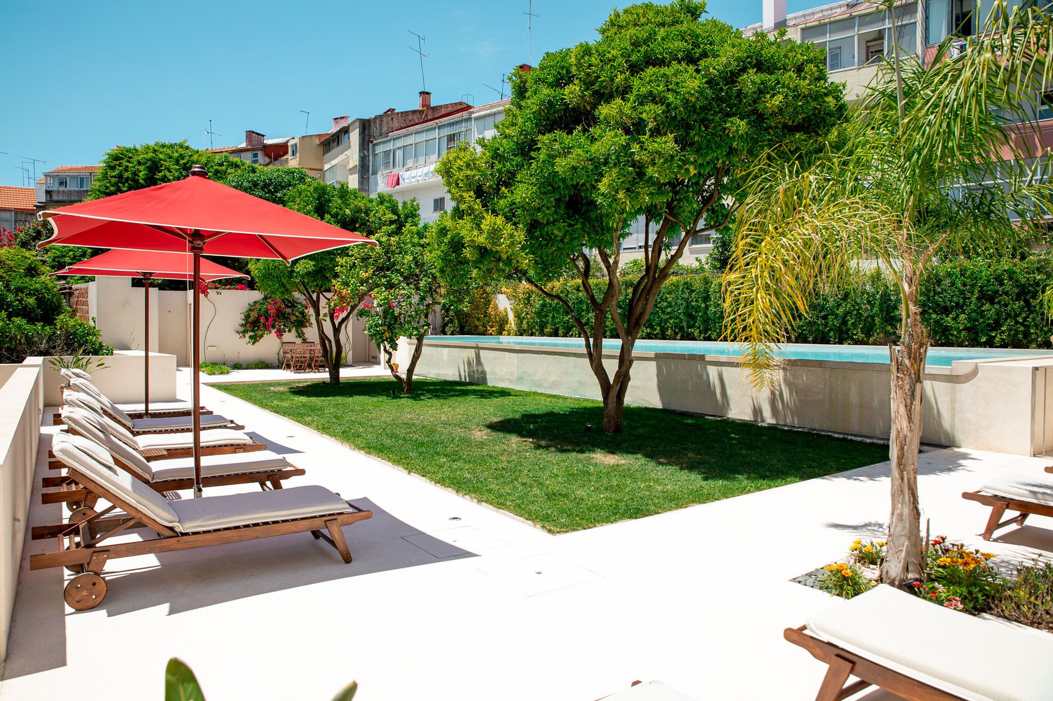 Uma área ao lado da piscina com espreguiçadeiras e um guarda-sol vermelho, cercada por vegetação e um edifício ao fundo.