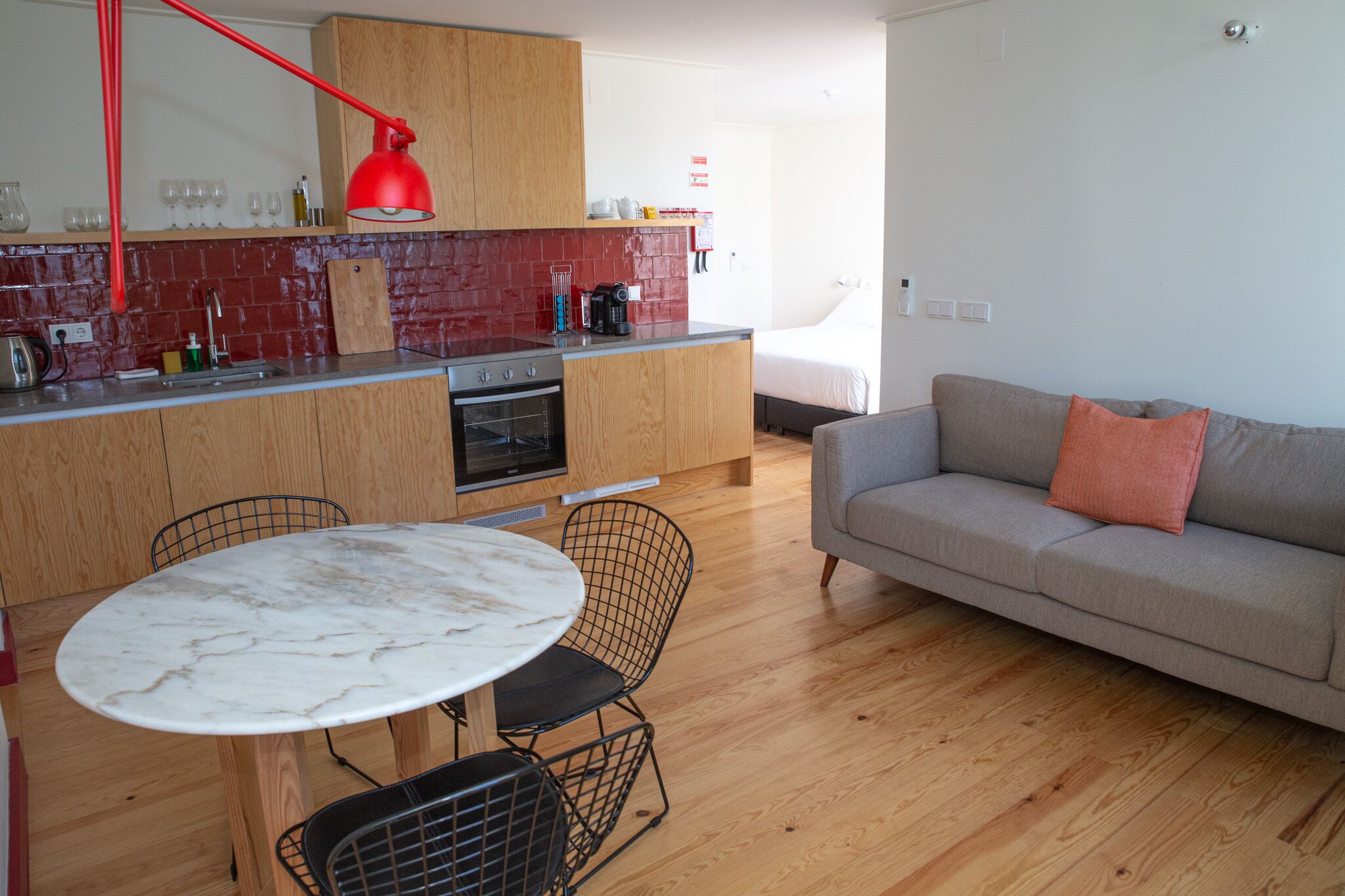 A compact living space with a kitchenette, a dining area with a marble table, and a grey sofa with an orange pillow.