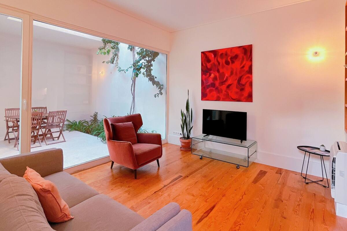A modern living room with a sofa, a red armchair, a TV on a glass stand, and a dining area with chairs and a table.