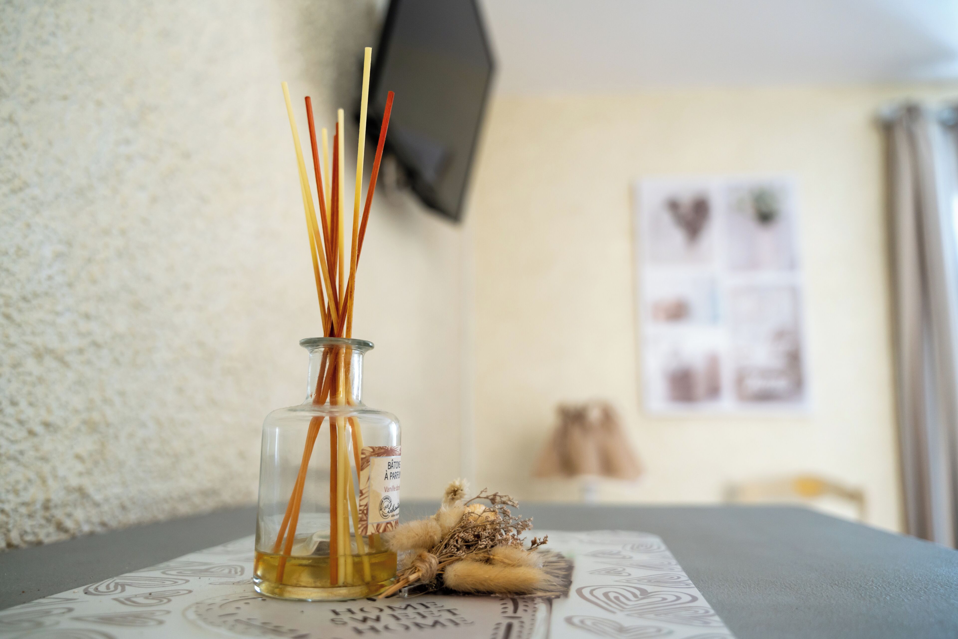 Un flacon en verre contenant des tiges de roseau et des fleurs séchées, posé sur une table recouverte d’une nappe à motifs.