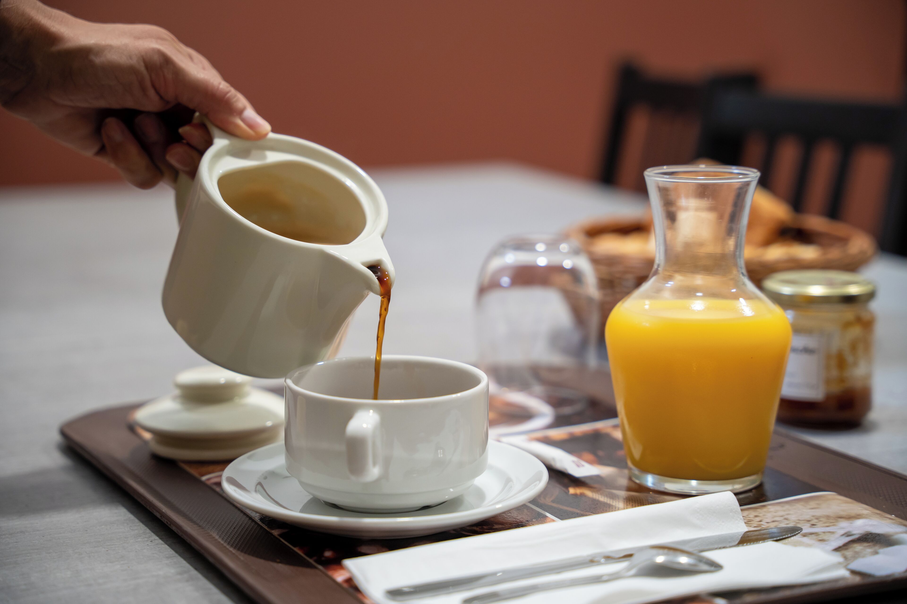 A hand pouring coffee into a cup, a glass of orange juice, and a jar of jam on a tray.