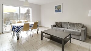 Een moderne woonkamer met een grijze bank, een eettafel met stoelen en een groot raam.