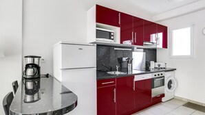 Een kleine keuken met rode kastjes, een witte koelkast, een magnetron, een koffiezetapparaat en een wasmachine.