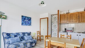 A cozy room with a blue patterned sofa, a wooden dining table with chairs, a kitchen area with wooden cabinets, and a framed abstract painting on the wall.