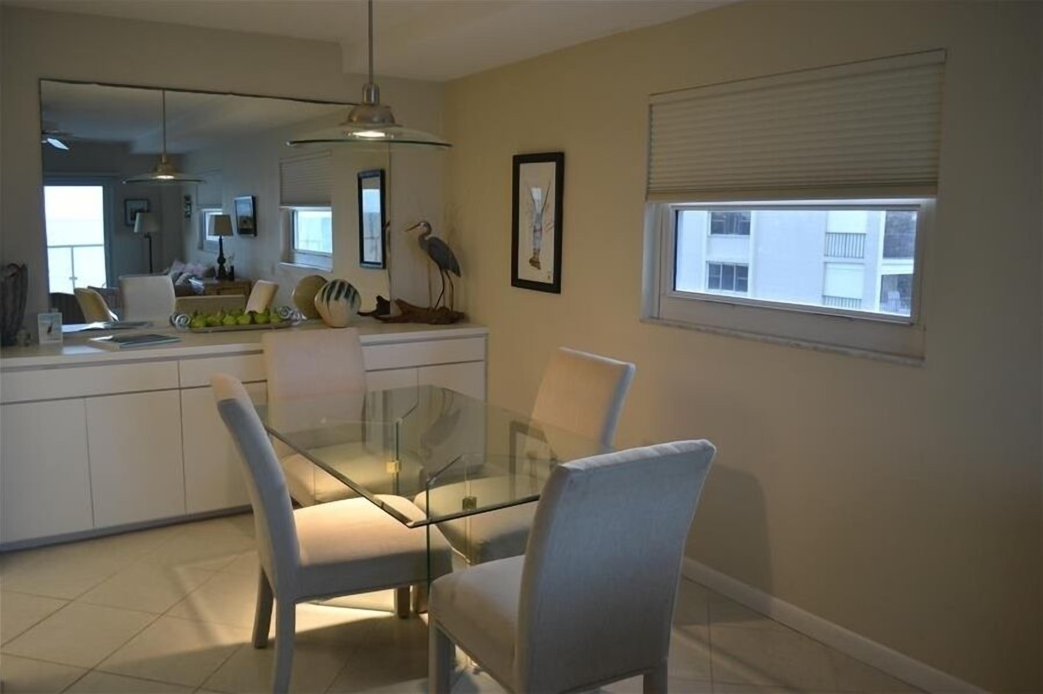A modern dining area with a glass table and chairs, a kitchen in the background, a framed artwork on the wall, and a window with blinds.