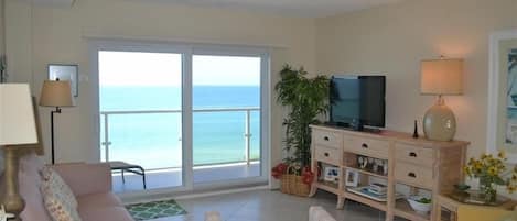 A living room with a sofa, a striped chair, a television on a wooden cabinet, a lamp, and a view of the sea through large glass doors.