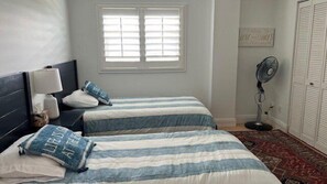 A bedroom with two beds, a nightstand, a bedside lamp, a fan, and a window with blinds.