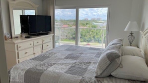 A bedroom with a large bed, a television on a dresser, a window with a view of buildings, and a lamp on the nightstand. - Beachcomber unit 603 (Longboat Key)