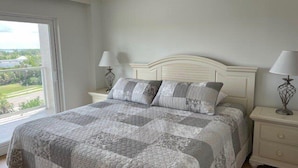 A bedroom with a bed, two nightstands, two lamps, and a window with a view of greenery. - Beachcomber unit 603 (Longboat Key)