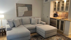 A modern living room with a grey sectional sofa, a coffee maker, and a kitchen area with cabinets and a countertop. - Beachcomber unit 603 (Longboat Key)