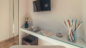 Een moderne kamer met een glazen bureau, een flatscreen televisie, een vaas met bloemen, een dienblad met koekjes en een theepot, en een glazen pot met rietjes.