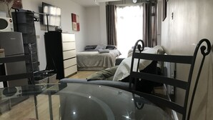 A compact hotel room with a glass table, a bed with a white blanket, a small desk with a lamp, and a TV on the wall. - Cultural quarter apartments (Leicester)