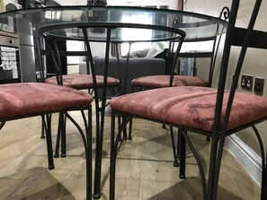 A dining area with a glass table surrounded by four chairs with cushioned seats. The floor is wooden, and there is a sofa in the background.