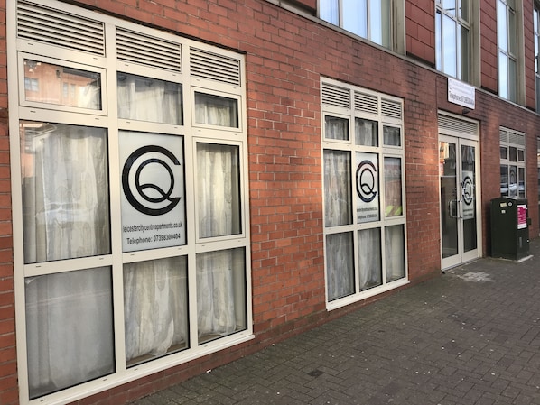 A building facade with multiple windows, some with curtains and others with blinds. There is a sign with a logo and contact information for a property management company.