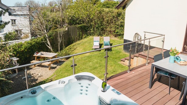 En terrasse med jacuzzi, et bord og stole, med udsigt over en have med et hegn og græsplæne.