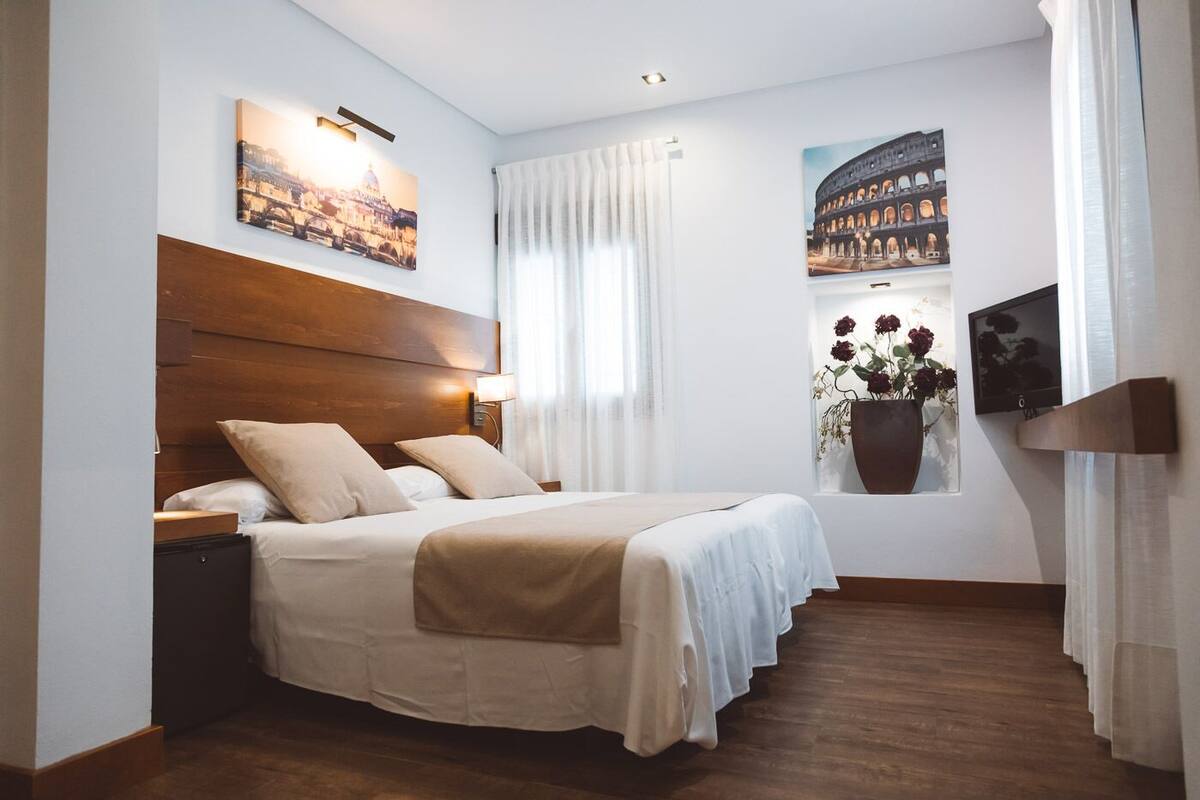 A hotel room with a large bed, a wooden headboard, a nightstand, a wall-mounted TV, and a window with a view of a building.