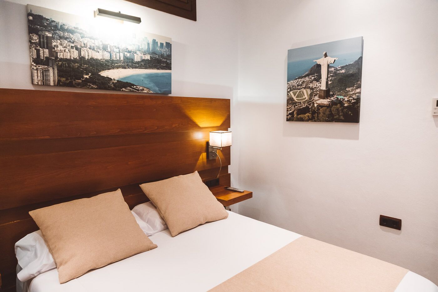 Een hotelkamer met een houten hoofdeinde, een bed met twee kussens, een nachtkastje met een lamp en twee ingelijste foto’s aan de muur.