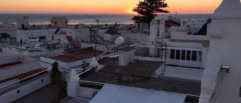 Terraza en la azotea con vistas a la puesta de sol sobre el mar, edificios blancos y un árbol.