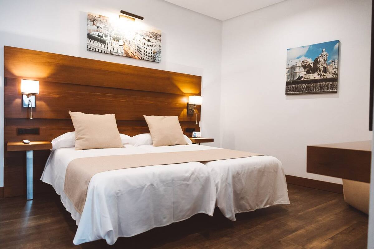 A hotel room with a wooden headboard, two bedside tables with lamps, a bed with white linens, and artwork on the walls.
