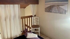 Une chambre avec un plafond en bois, une chaise blanche, un lit recouvert d’une couverture violette et un tableau représentant une scène de plage.