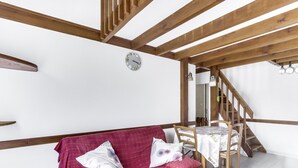 Une pièce chaleureuse avec un plafond en bois, un canapé à carreaux rouges agrémenté de coussins, une petite table recouverte d’une nappe blanche et une horloge au mur.