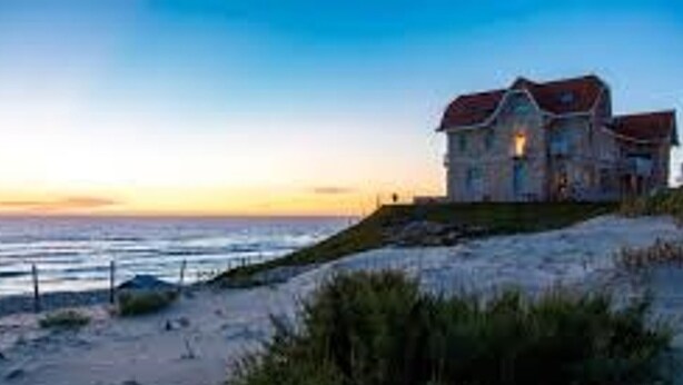 Une maison située sur la côte, avec un toit rouge, qui donne sur une plage de sable et offre une vue sur l’océan.