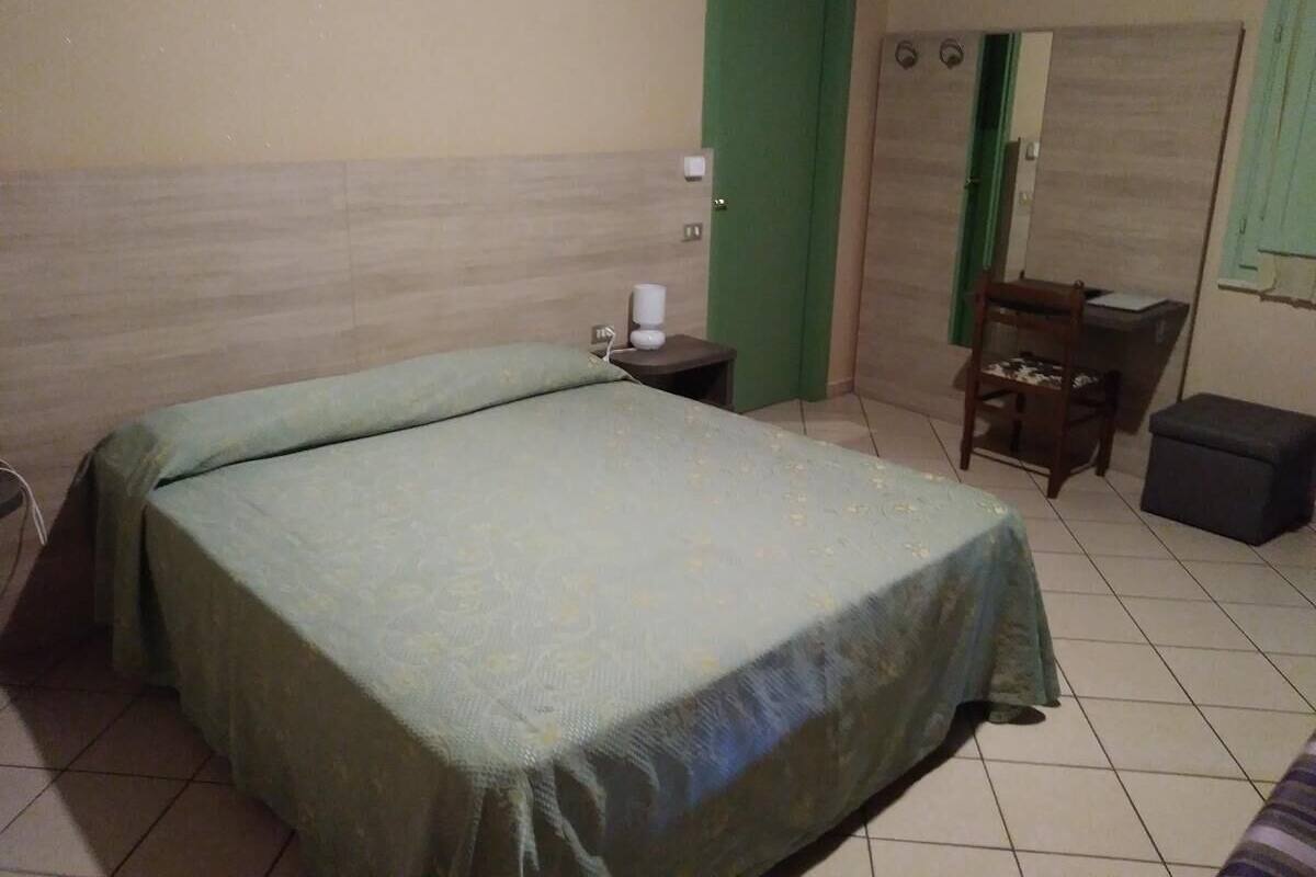 A single-bed room with a green bedspread, a bedside table with a lamp, a chair, and a small table with a chair.