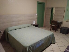 Ein Einzelzimmer mit einem Bett mit grüner Tagesdecke, einem Nachttisch mit Lampe, einem Sessel und einem kleinen Tisch mit Stuhl.