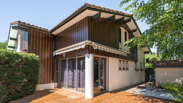 Una casa de dos pisos con un balcón de madera, grandes puertas corredizas de vidrio y techo de tejas.