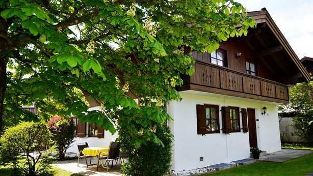 Une maison en bois à deux étages, dotée d’un balcon, d’un petit jardin et d’une table avec des chaises à l’extérieur.