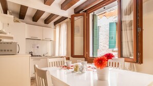 A kitchen with white cabinets, a white countertop, and a dining table with chairs. There is a window with green shutters and a vase with flowers on the table. - San Marco Joy (Venice)
