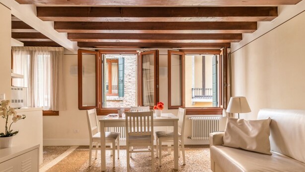A room with a wooden ceiling, a white sofa, a dining table with chairs, and a window with a view of a building. - San Marco Joy (Venice)