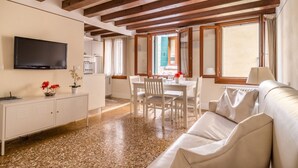 A spacious living area with a white sofa, a dining table with chairs, a TV mounted on the wall, and a kitchen in the background. - San Marco Joy (Venice)