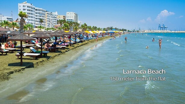 A beach with sun loungers and umbrellas, a clear sky, and buildings in the background. - Aladdin's Hostel (Larnaca)
