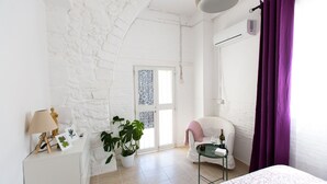 A room with a white brick wall, a white door with a window, a white chair, a small table with a bottle, and a bed with a floral bedspread. - Aladdin's Hostel (Larnaca)
