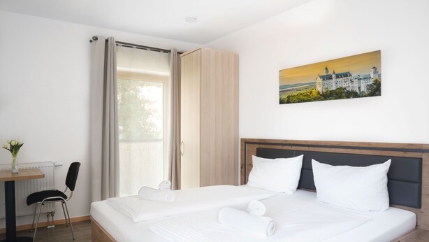 A hotel room with a large bed, a desk with a chair, a window with curtains, and a painting on the wall.
