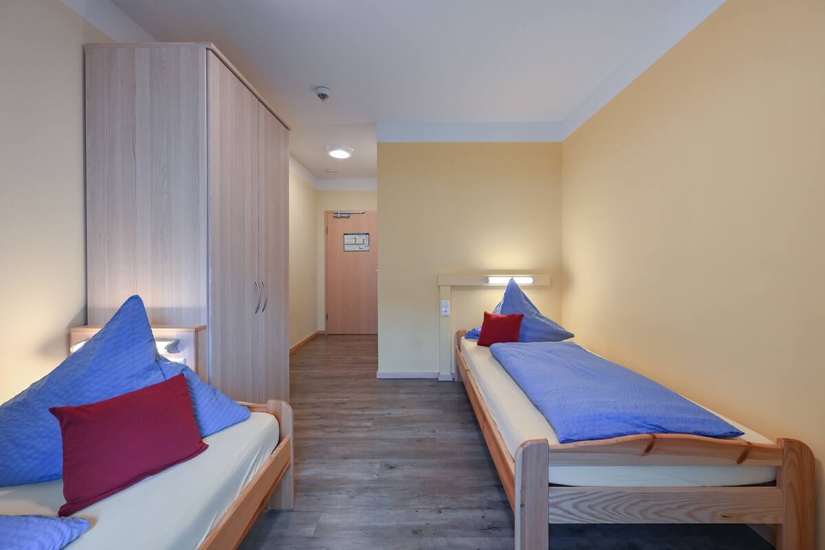 A room with two single beds, each with a blue mattress and a red pillow. There is a wooden wardrobe on one side and a door at the end of the room.